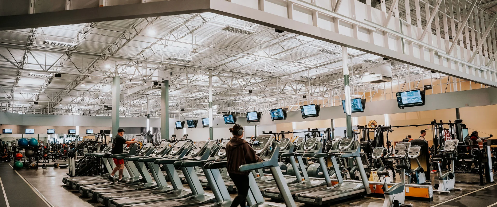 Cardio area at Gold's Gym Smyrna, Tennessee, featuring rows of treadmills and stationary bikes with individuals exercising. Overhead TVs and high ceilings with industrial beams are visible in the background.
