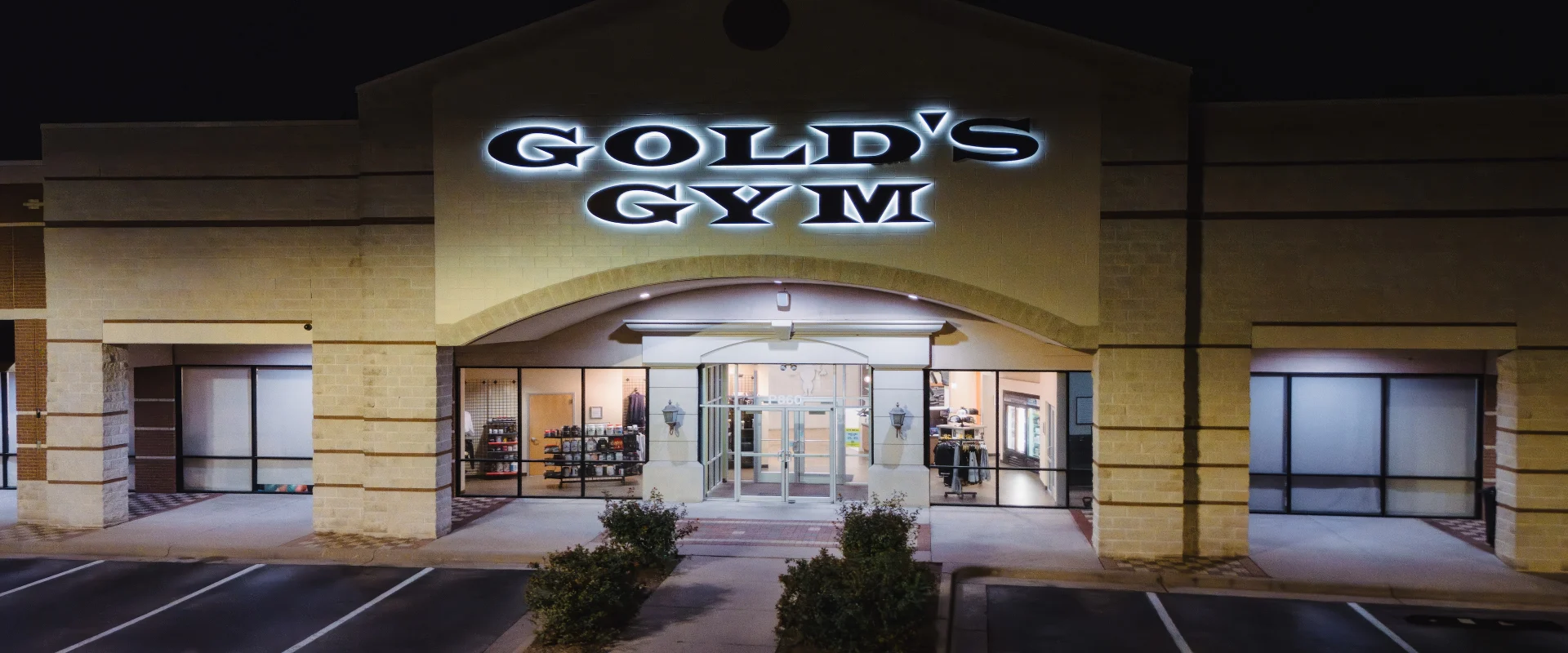 Exterior view of Gold’s Gym in Austin (Westlake), captured at night with the bold, illuminated Gold’s Gym sign glowing above the entrance. The modern facade features large glass doors, flanked by windows revealing the well-lit interior. The front walkway is neatly landscaped, with an empty parking lot in the foreground.