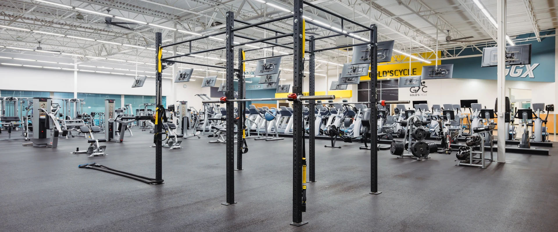 Functional training area at Gold’s Gym in Texas (N. Round Rock), featuring a multi-station rig with pull-up bars, suspension straps, and barbell holders. The surrounding space includes resistance machines, cardio equipment, and free weights. Overhead, mounted TVs face the cardio machines, and the Gold’s Cycle and GGX areas are visible in the background. The flooring is rubberized for impact absorption, with high ceilings and bright overhead lighting.