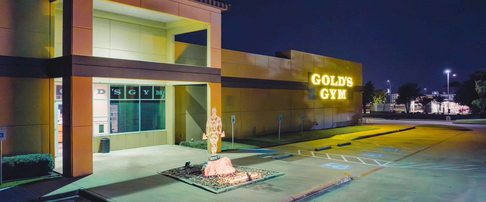 Gold's Gym College Station exterior with illuminated Gold's Gym logo and iconic bodybuilder silhouette centered and brightly lit at the entrance