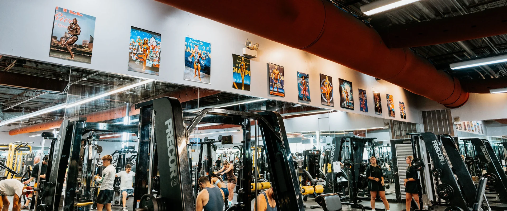 Members exercising in the plate-loaded equipment area at Gold’s Gym Bellevue, Nashville, Tennessee. The gym features machines and members engaged in various exercises. The mirrored wall reflects the equipment and gym-goers, while a series of bodybuilding competition posters and awards line the upper wall. Overhead, industrial-style lighting and a large red ceiling duct add to the gym's dynamic ambiance.