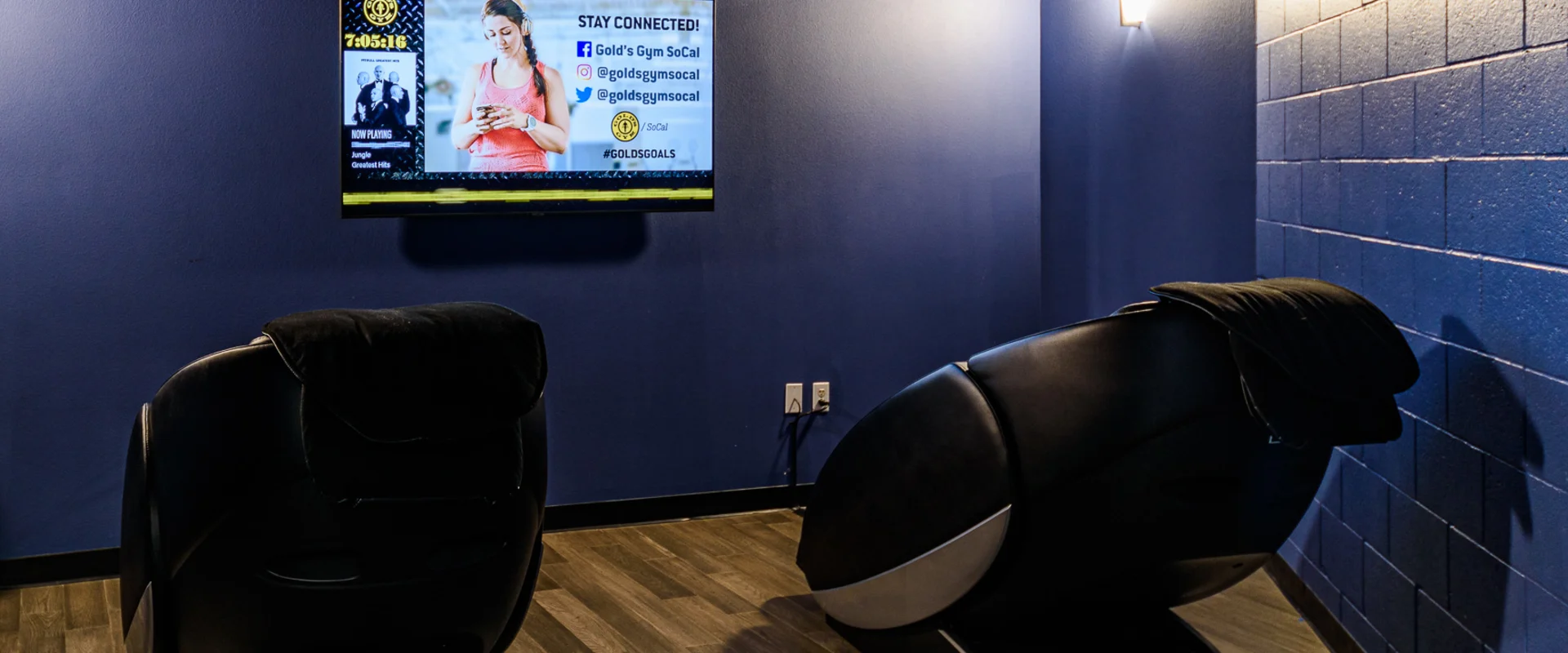 A dimly lit recovery room featuring two black HydroMassage chairs with built-in controls. A large screen on the blue accent wall displays Gold’s Gym promotional content, while a soft wall light provides ambient lighting. The floor is covered in dark wood laminate, enhancing the relaxation space.