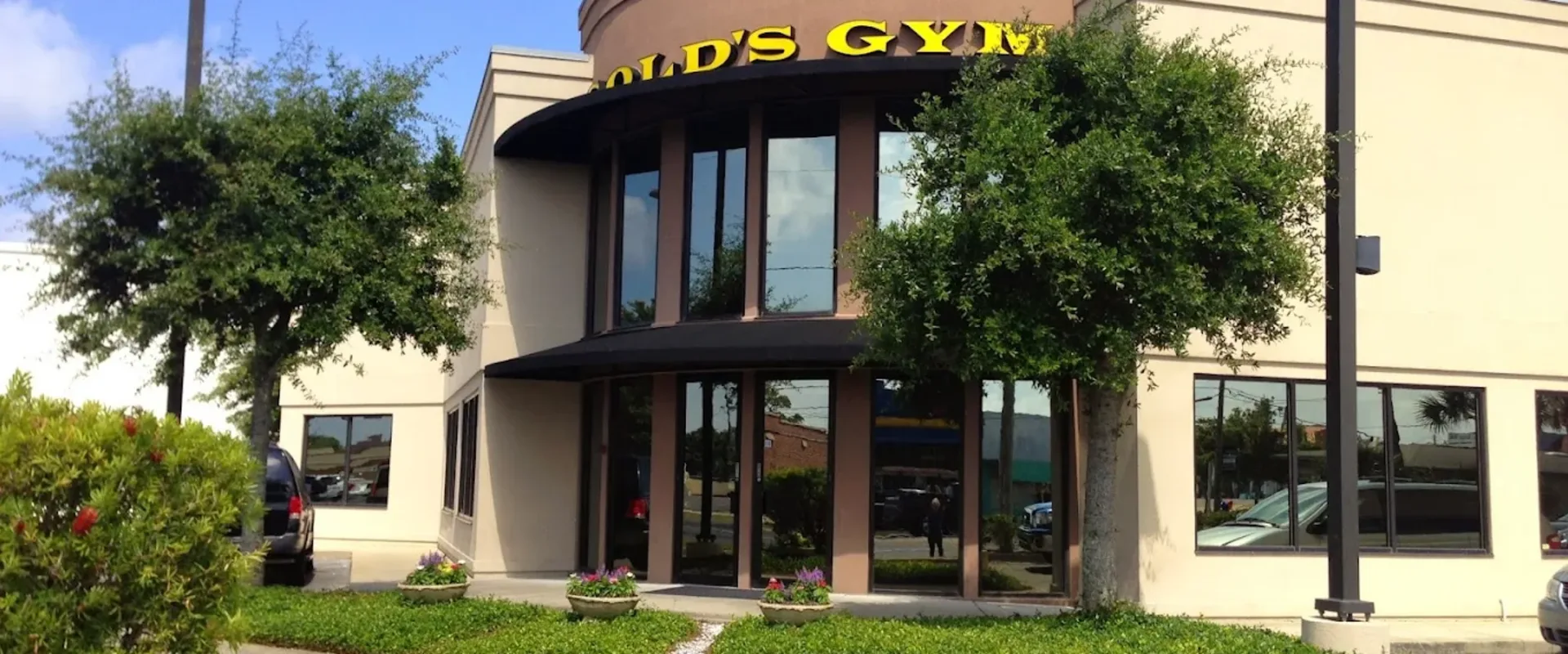 Exterior of Gold’s Gym Fort Walton Beach with curved glass entrance, yellow signage, and landscaping.