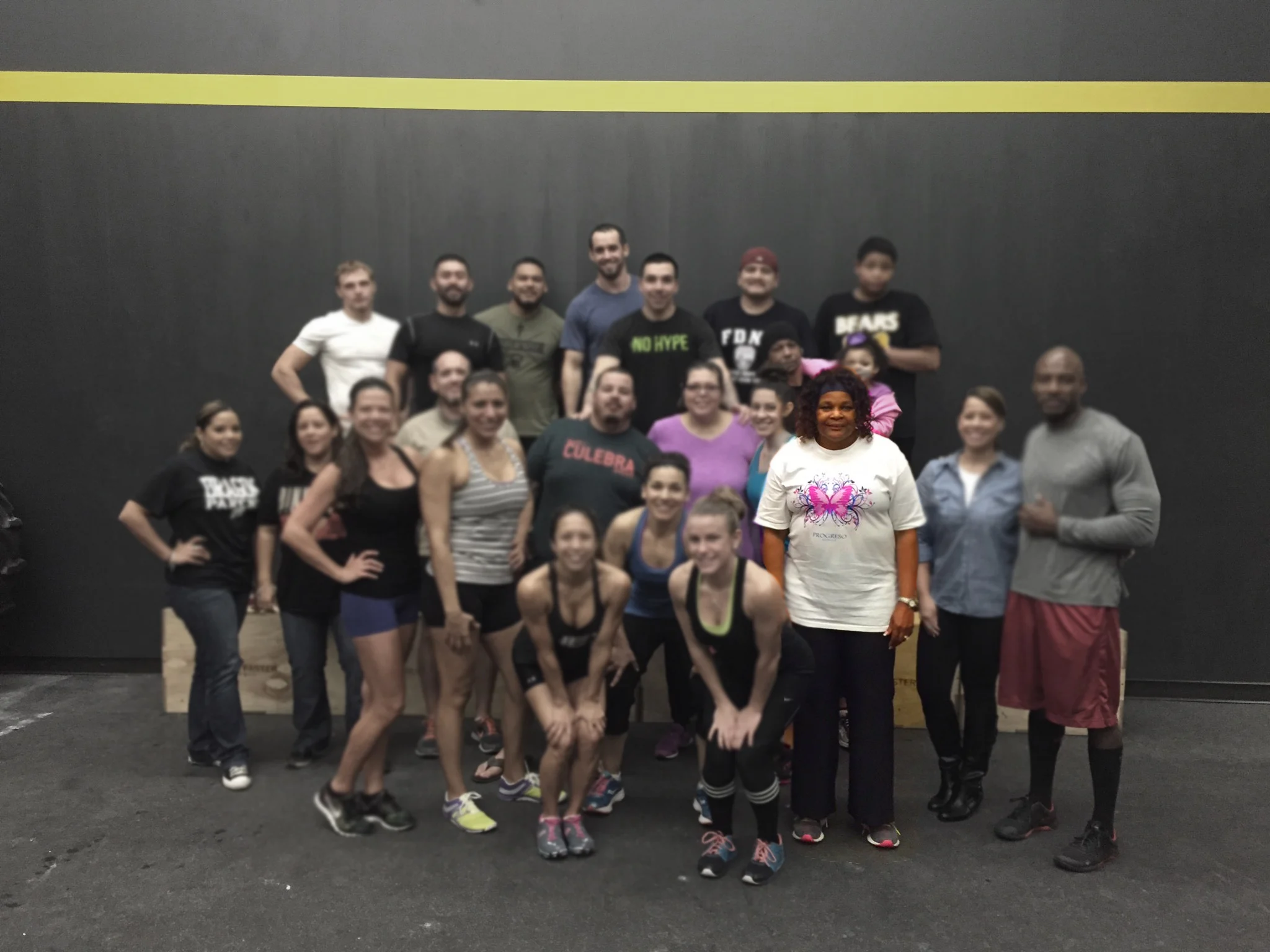 A group of people, both men and women, are gathered together in what appears to be a fitness or exercise studio, with a gray wall and a yellow line in the background.