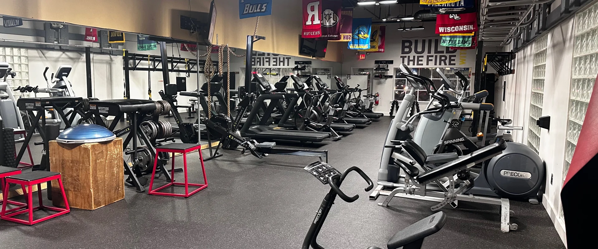 Gold's Gym Bridgewater circuit training area featuring various strength and conditioning equipment. Plyometric boxes, stability balls, resistance machines, and cardio equipment are arranged for functional workouts. Overhead, college banners hang from the ceiling. The mirrored wall reflects the training space, and battle ropes are mounted for high-intensity exercises. The area is designed for dynamic circuit workouts with a mix of weight and endurance training stations.