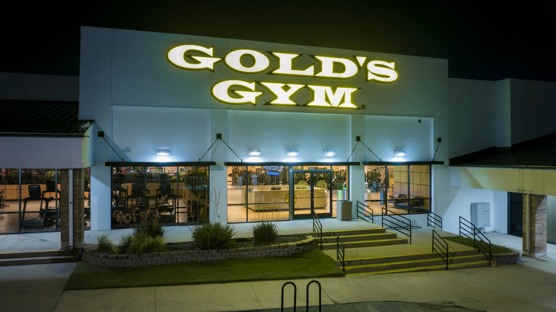 Nighttime exterior of Gold’s Gym Highland Meadows with a brightly illuminated gold sign above the entrance. The modern facade features large glass doors and windows showcasing the interior. The surrounding area includes landscaped greenery, accessible ramps, and a parking lot in the foreground, all softly lit by overhead lights.