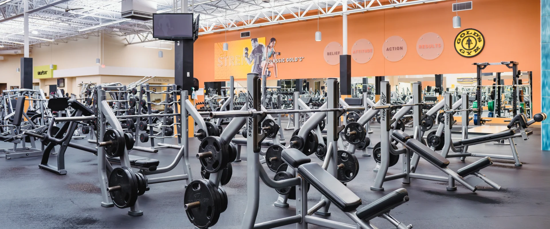 Bright, spacious gym interior with rows of weightlifting equipment and benches. Orange wall with motivational words enhances the energetic atmosphere.