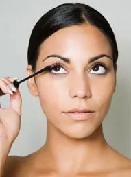 A woman with dark hair is applying makeup, specifically eyeliner, against a plain gray background.
