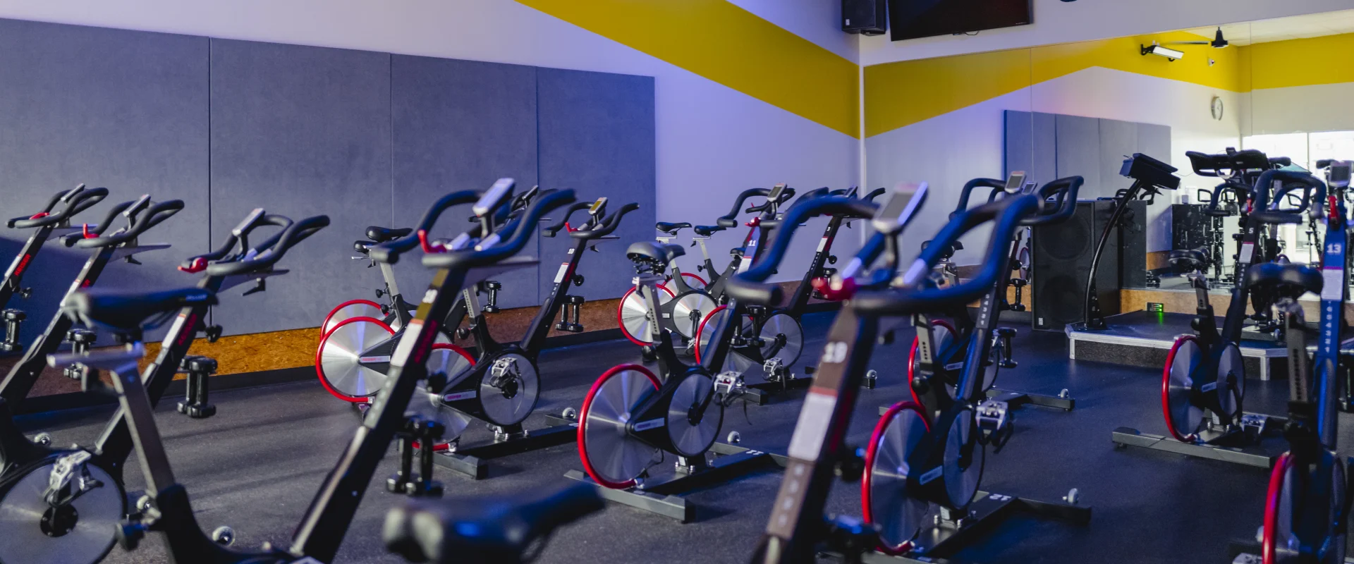Cycle studio at Gold's Gym College Station (Tower Point), featuring rows of stationary bikes, mirrored walls, padded acoustic panels, and bright yellow accents.