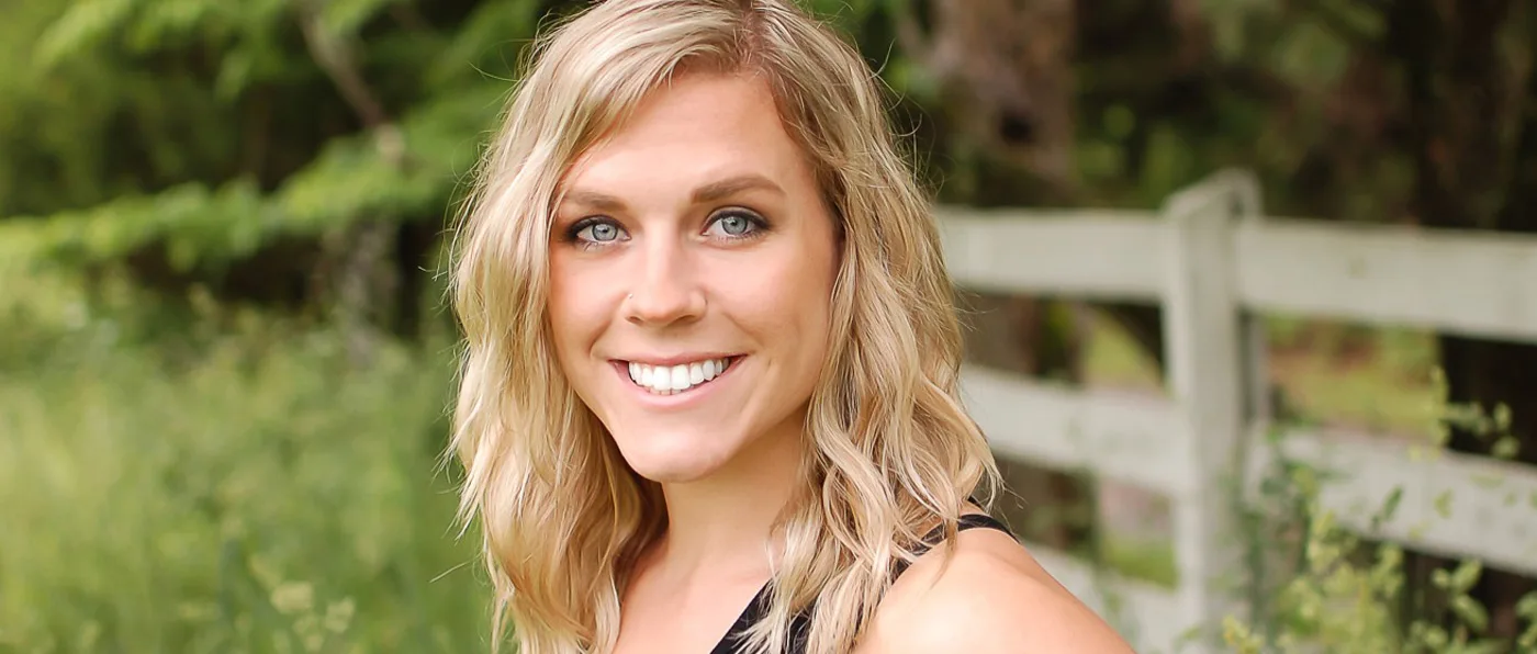 A smiling young woman with long blonde hair stands in a lush, green outdoor setting, with a wooden fence visible in the background.
