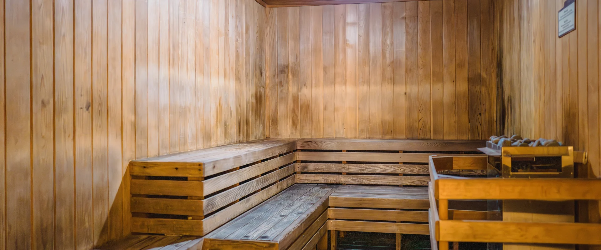 Spacious wooden sauna interior with warm lighting. Wooden benches line the walls, and a heater with stones sits on the right, creating a cozy, relaxing ambiance.