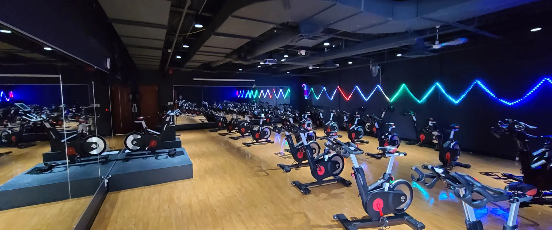 Indoor cycling studio at Gold’s Gym Vertu featuring rows of black-and-red stationary bikes on wood flooring, mirrored walls, and colorful zigzag LED lighting on black walls.