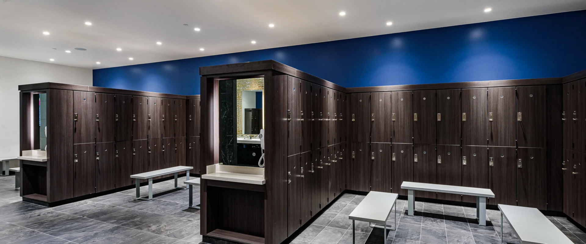 The locker room at Gold’s Gym Cerritos features dark wood lockers with silver handles, arranged around a central vanity area with built-in mirrors and countertops. White benches are placed throughout the space, and the floor is covered in large gray tiles. Recessed lighting illuminates the area, and a deep blue accent wall adds contrast.