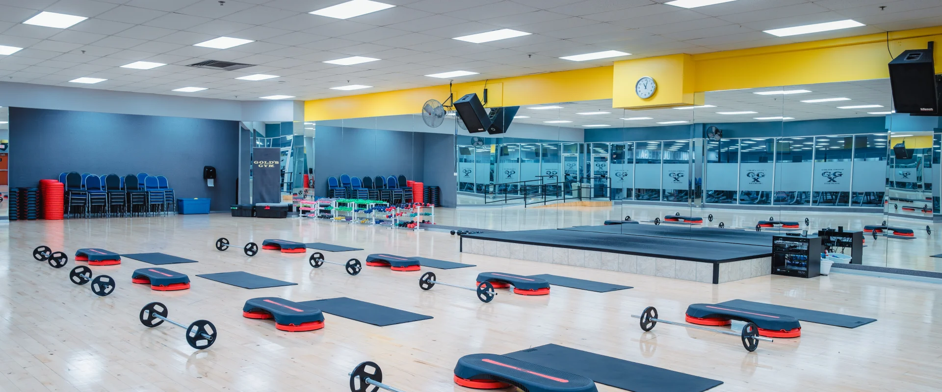 Group exercise studio at Gold’s Gym in Austin (South Central), featuring a spacious hardwood floor with workout stations set up with mats, barbells, and step platforms. The room includes a raised instructor stage, mirrored walls for form checks, and bright overhead lighting. Stacked equipment and chairs are neatly organized along the perimeter, with large windows providing natural light and views of the gym floor.