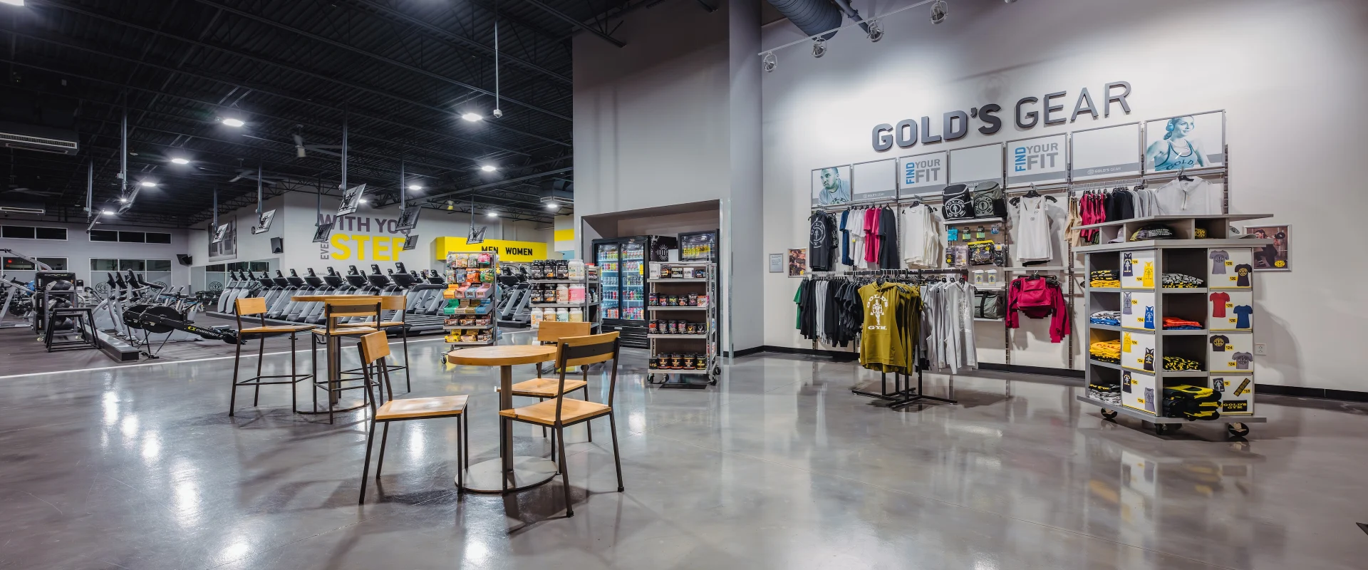 The Pro Shop area features racks displaying branded apparel, including t-shirts, hoodies, and athletic gear. Several shelves are stocked with supplements, snacks, and fitness accessories. Small round tables with chairs provide a casual seating area, positioned near the cardio equipment section in the background.