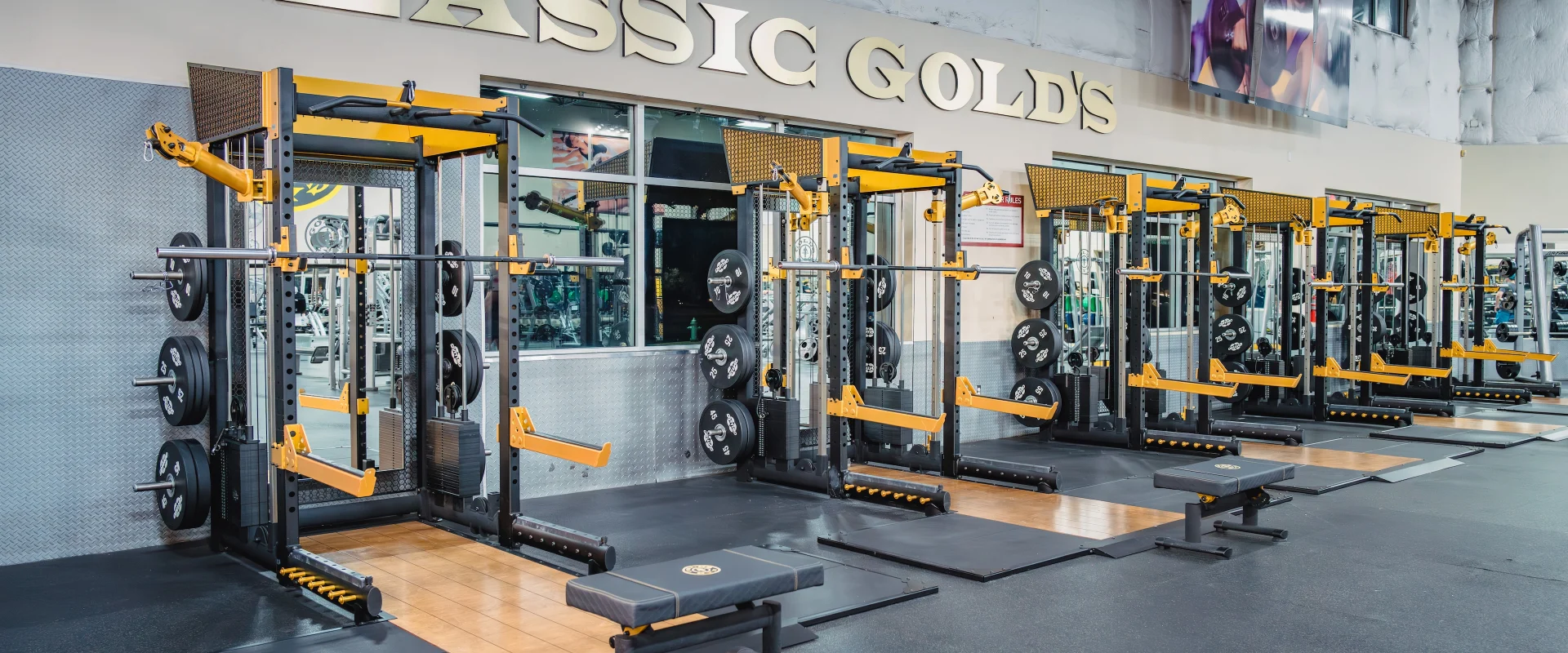 The Olympic lifting platforms at Gold’s Gym in Austin (South) feature multiple lifting stations with black and yellow power racks, each equipped with barbells and neatly stacked weight plates. The platforms have a combination of wooden centers and black rubber edges, providing stability and shock absorption. A padded bench is positioned in front of one platform, and mirrors behind the racks reflect the gym’s interior. The wall above displays bold “CLASSIC GOLD’S” signage, adding a signature touch to the space.