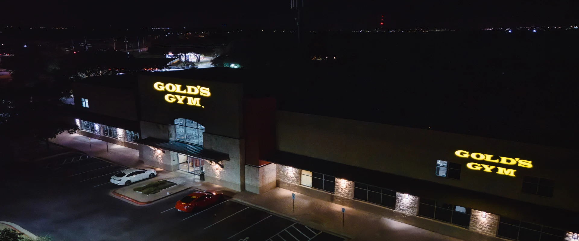 The exterior of Gold’s Gym in Austin (South) is illuminated at night, with two bright, yellow-lit “GOLD’S GYM” signs mounted on the building’s facade. The entrance features large glass doors framed by stone accents and a metal awning. The well-lit parking lot in front has a few cars parked near the entrance, with marked spaces, including designated handicapped spots. The surrounding area fades into darkness, with distant city lights visible on the horizon.