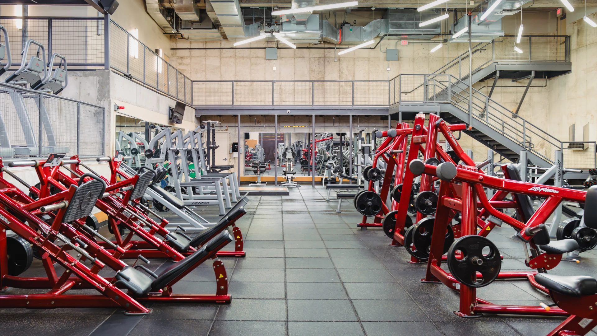 Strength training area at Gold's Gym Austin (Downtown) featuring plate-loaded machines and benches with bold red frames. The industrial-style space includes concrete walls, modern overhead lighting, and rubber flooring designed for durability and comfort.