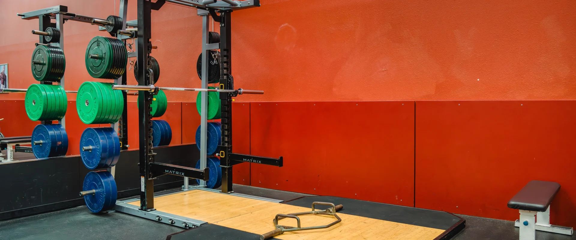 A gym with a squat rack, colorful weight plates, and a barbell against a red wall. A bench and a rope are on the black and wooden floor.