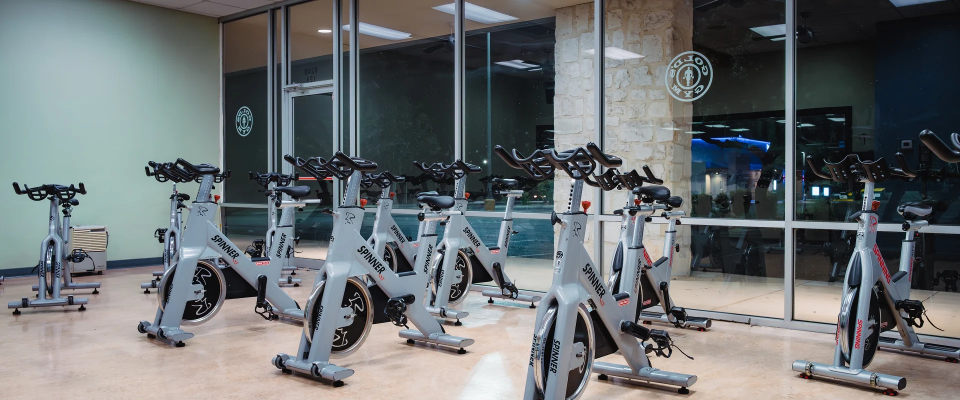 A row of stationary spin bikes in a well-lit gym with large windows reflecting interior light. The room has a calm and orderly atmosphere.