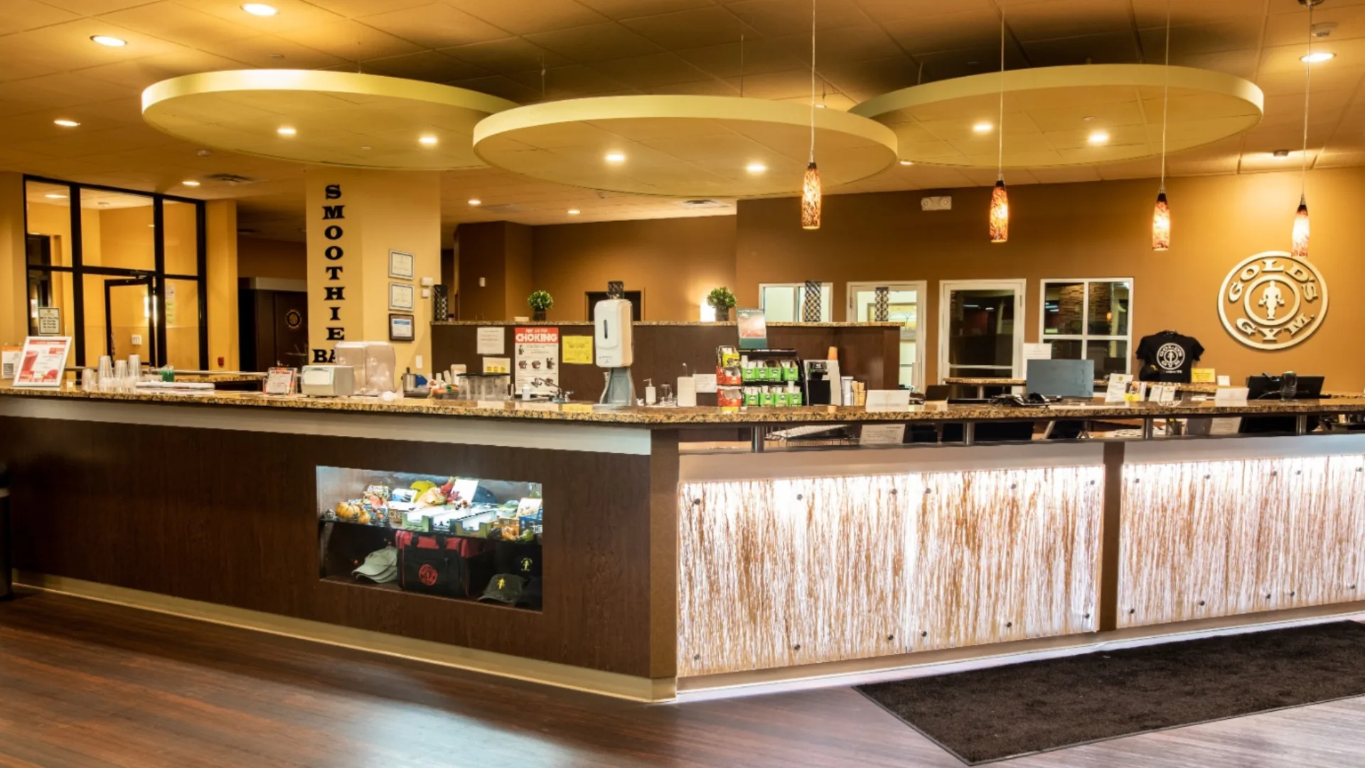 Smoothie bar at Gold’s Gym in East Northport, New York, featuring a granite countertop with a display of supplements, protein snacks, and branded merchandise. The bar includes a hand sanitizer station, blenders, and stacked cups for quick service. Pendant lights and large circular ceiling fixtures provide warm lighting, while the Gold’s Gym logo is prominently displayed on the back wall.