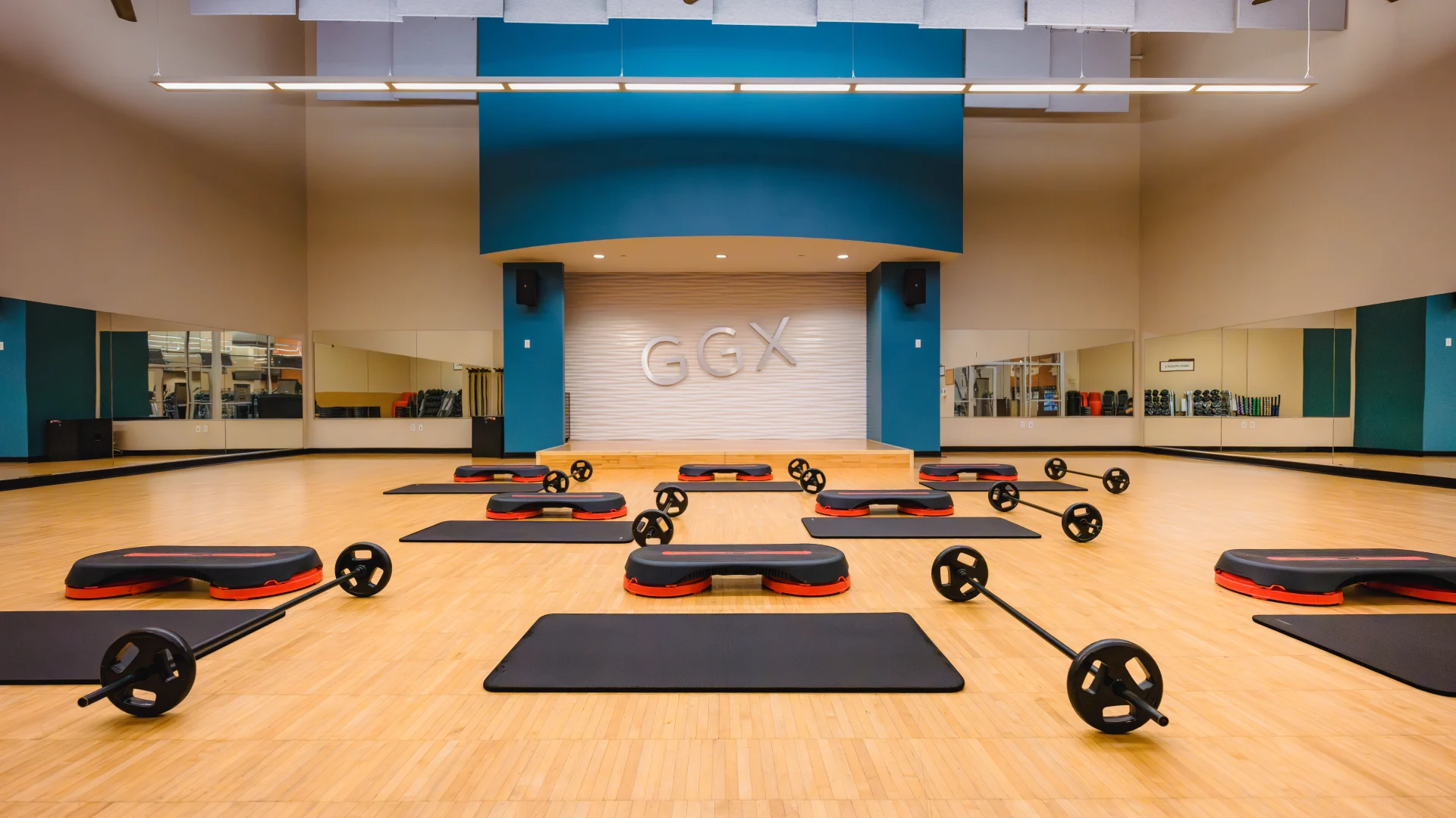 Group exercise studio at Gold’s Gym Austin (Anderson Arbor) featuring a wooden floor, step platforms, mats, and barbells arranged in rows. A stage with the “GGX” logo is centered on the teal and white accent wall, with large mirrors lining the sides of the room.