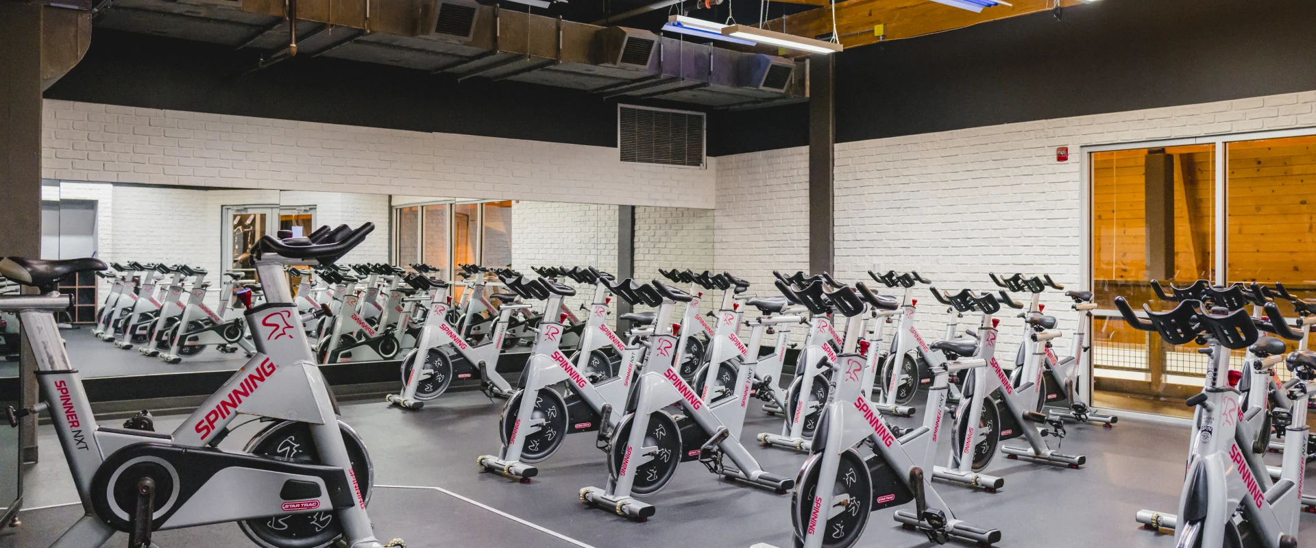 A group cycling studio with rows of stationary bikes, a central instructor platform, mirrored walls, and a wooden ceiling with bright lighting.