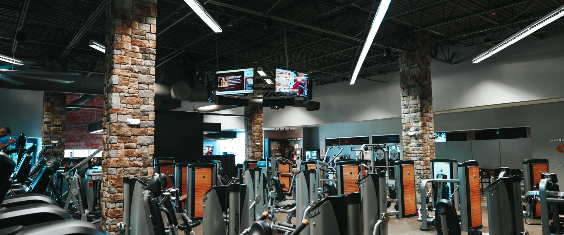 Strength training area at Gold's Gym Rock Hill, filled with rows of resistance machines and plate-loaded equipment. The industrial-style space features exposed ductwork, stone pillar accents, and high ceilings, creating a rugged yet modern workout environment.