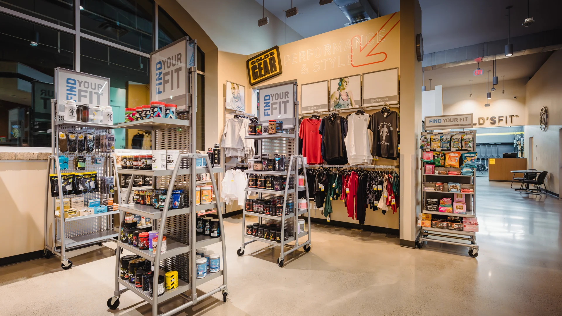 Pro Shop at Gold's Gym Austin (Downtown) showcasing shelves stocked with workout supplements, protein powders, energy bars, and fitness accessories. The display also includes racks of gym apparel such as hoodies, tank tops, and leggings. Bright lighting highlights the clean, modern retail space with promotional signs and a polished concrete floor.