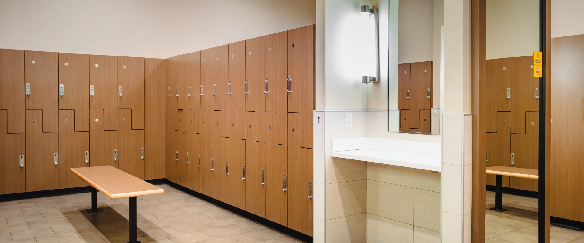 Spacious locker room with wooden lockers and a bench. A large mirror is beside a counter with two wall lights, creating a clean, organized feel.