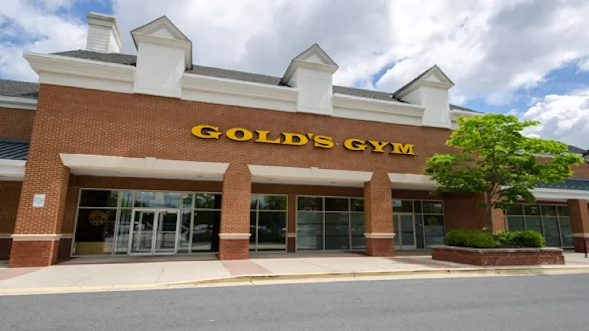 Exterior of Gold’s Gym Bowie with main entrance, brick facade, and yellow logo sign on a sunny day.