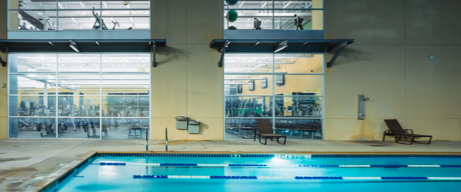 Indoor swimming pool with blue lane dividers, reflecting gym windows in the background. The area feels clean and inviting, with lounge chairs nearby.