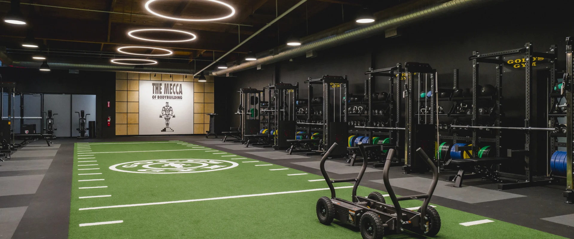 A turf training area at Gold's Gym featuring a sled, functional equipment racks, and a wall with 'The Mecca of Bodybuilding' signage under modern lighting.