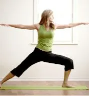 A woman in a green top and black pants is performing a yoga pose with her arms outstretched, standing on a yoga mat in a bright, open room with large windows.