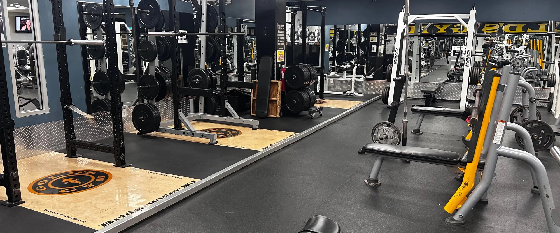 Olympic lifting platforms at Gold’s Gym New Jersey, Bridgewater, featuring multiple squat racks with attached barbell holders, bumper plates, and weightlifting stations. The platforms have a wooden center with the Gold’s Gym logo and are surrounded by black rubber for stability. The area has dark flooring, mirrored walls, and overhead lighting. Benches and additional weight machines are positioned nearby, with various strength training equipment visible in the background.