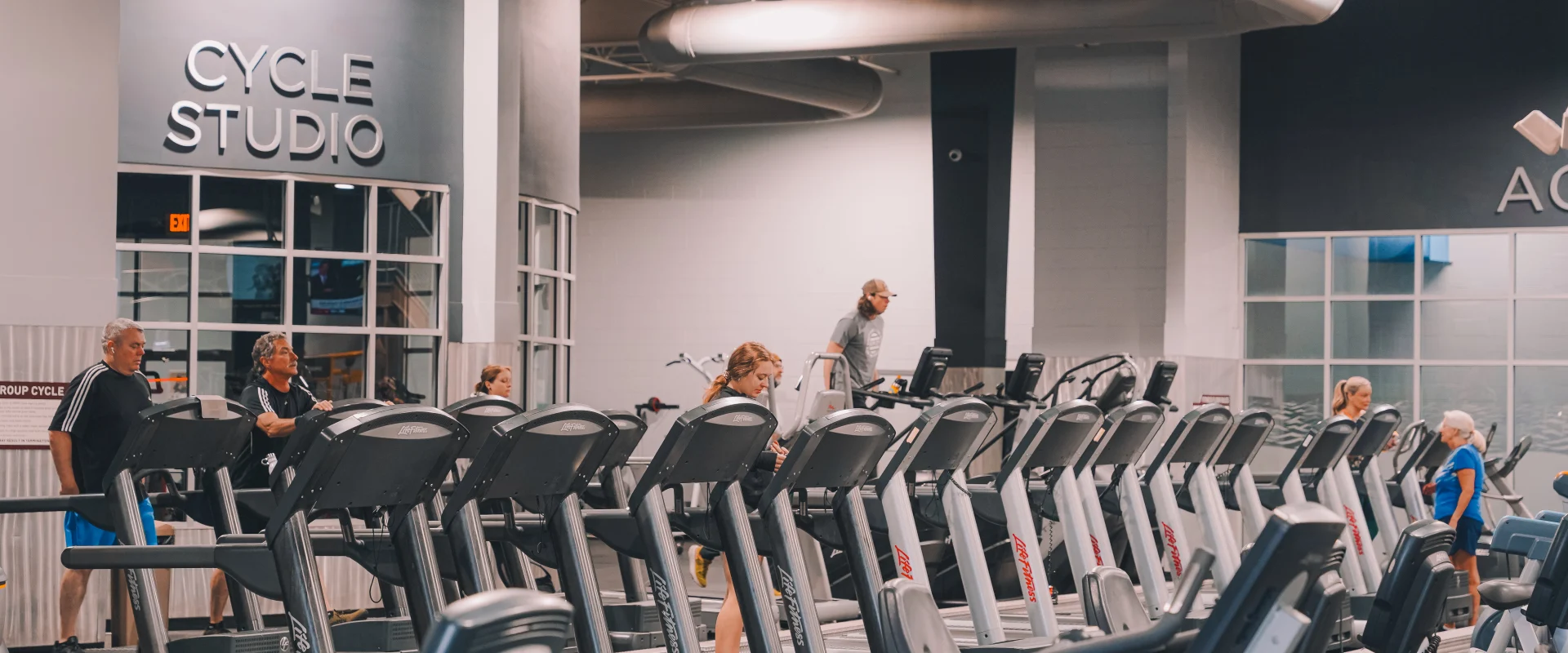 Members using treadmills in the cardio area at Gold’s Gym Farragut, Tennessee. The gym features a row of treadmills and stationary bikes, with members engaged in cardio exercises. The backdrop includes a Cycle Studio with glass-paneled walls and visible industrial-style ceiling ducts, creating a modern and functional workout space.