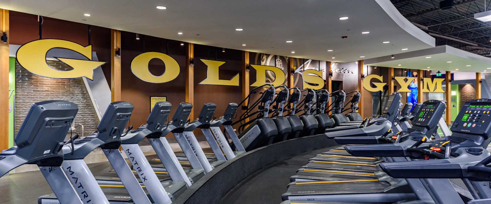 Rows of Matrix treadmills and elliptical machines face multiple mounted TVs, positioned on a curved black platform with overhead industrial lighting. The "Gold's Gym" logo in large yellow letters is displayed on the brown-paneled back wall.