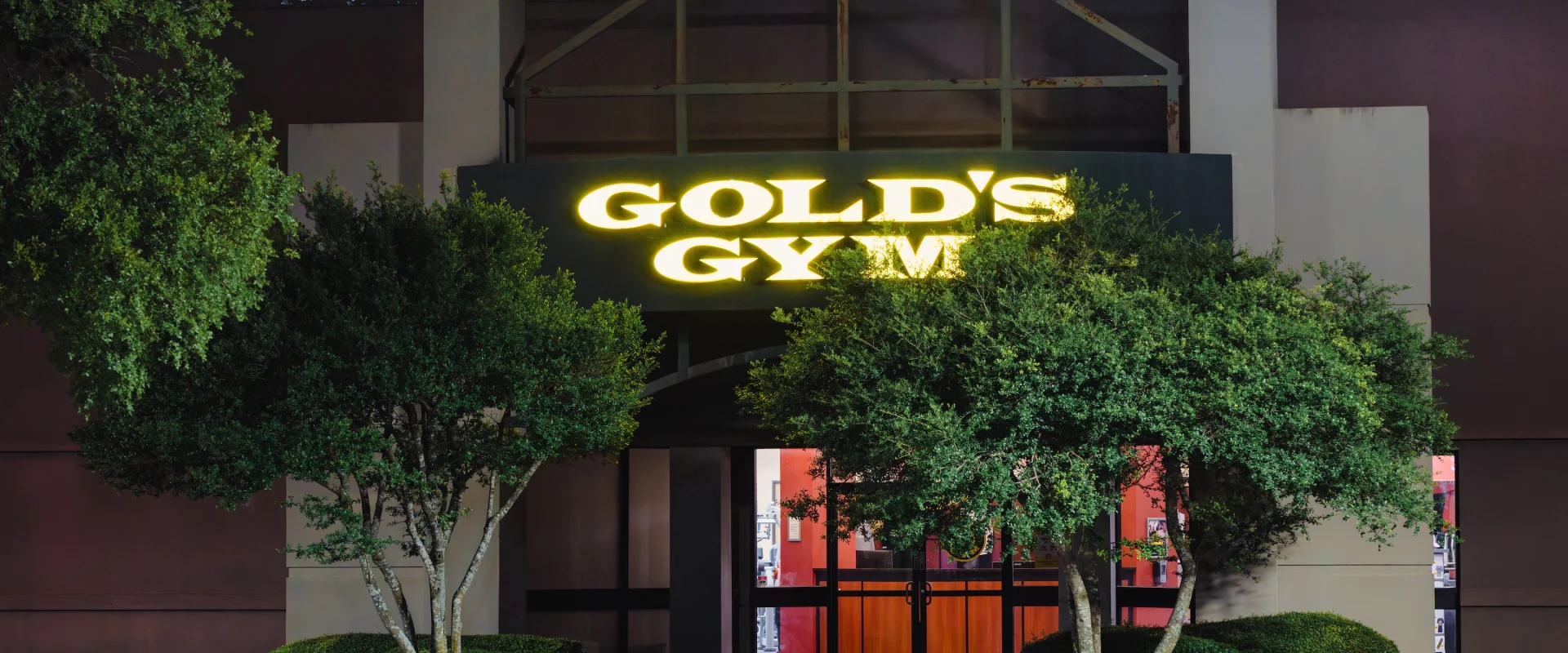 Exterior of a gym at night with a glowing "Gold's Gym" sign above the entrance, flanked by two large green trees and a well-lit entranceway.
