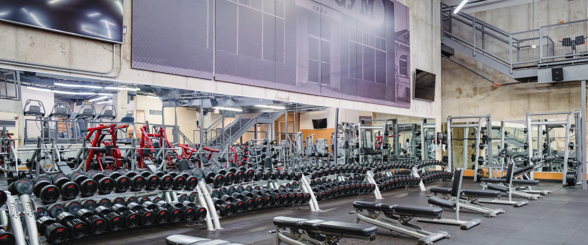Interior of Gold's Gym Austin (Downtown) with rows of dumbbells on racks, adjustable weight benches on rubber flooring, mirrored walls reflecting gym equipment, industrial-style light fixtures, a wall-mounted TV, and an old photo of Gold's Gym Venice displayed above the mirrors.