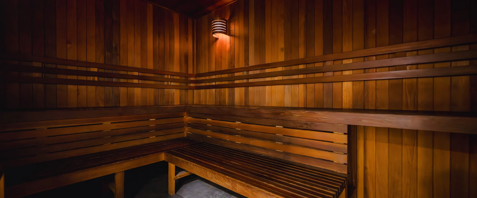 The sauna features wooden bench seating along the walls with horizontal slats, set against smooth, vertical wooden panels. Warm lighting fixtures mounted on the walls create a cozy ambiance, and tiled flooring lines the entry area.