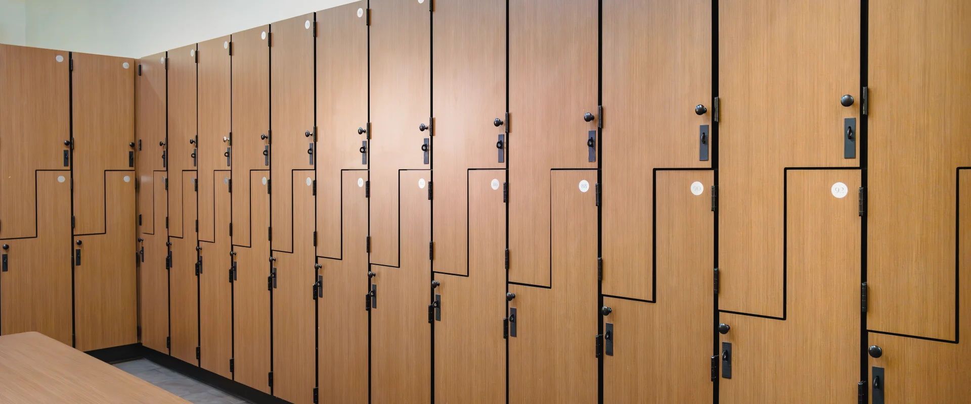 Locker room at Gold's Gym Austin (Downtown) featuring rows of wood-grain lockers with black hardware. A bench with a matching wood finish sits in front of the lockers, and the floor is tiled with light-colored ceramic.