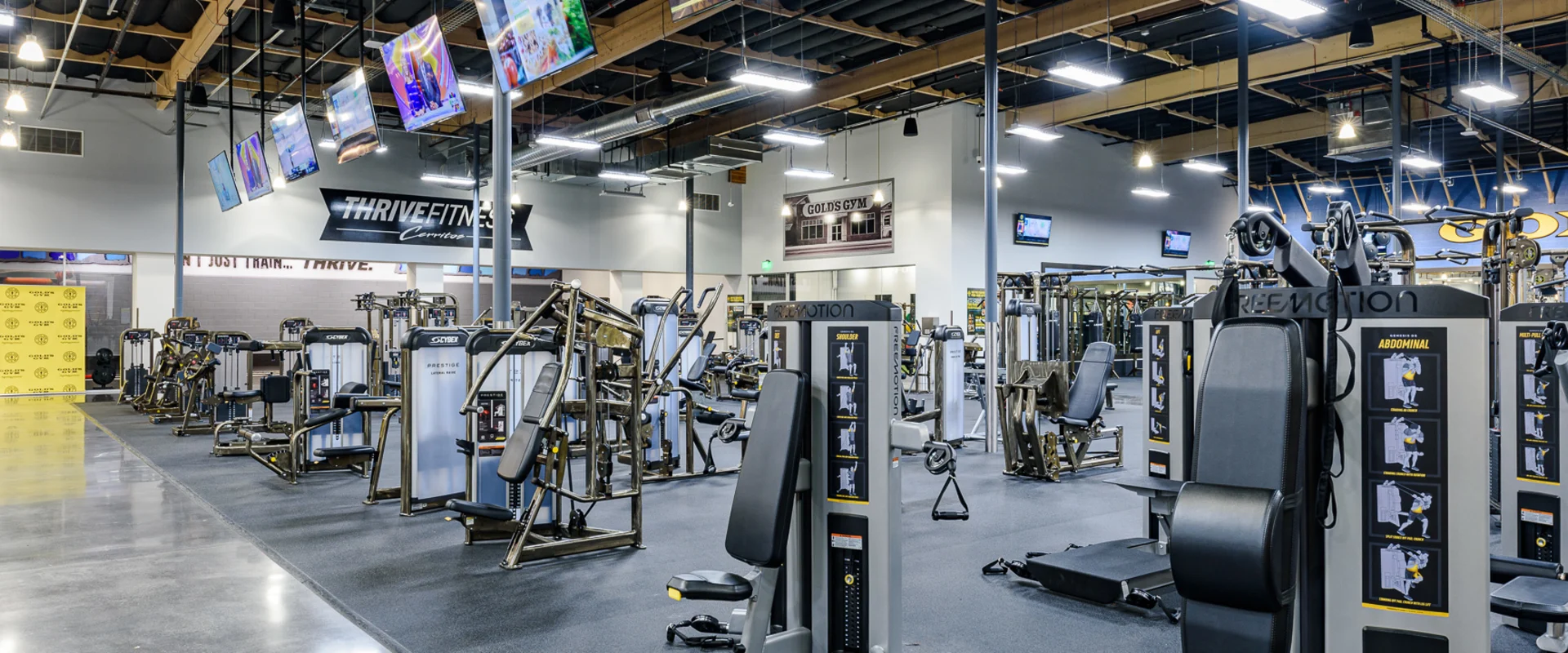 The weight machine area at Gold’s Gym Cerritos features rows of strength training machines with black padding and silver frames. Overhead TVs display fitness content, and a ‘Thrive Fitness Cerritos’ sign is visible in the background. The gym’s open layout includes black rubber flooring and exposed industrial-style lighting.