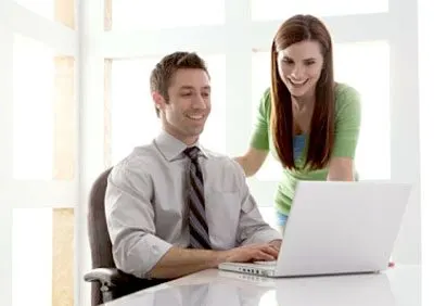 A young man in a suit is sitting at a desk, working on a laptop, while a smiling young woman stands next to him, appearing to be assisting or interacting with him.