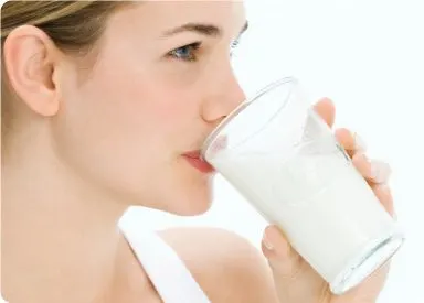 A woman with fair skin and light-colored hair is drinking from a glass of milk, her face in profile against a plain white background.