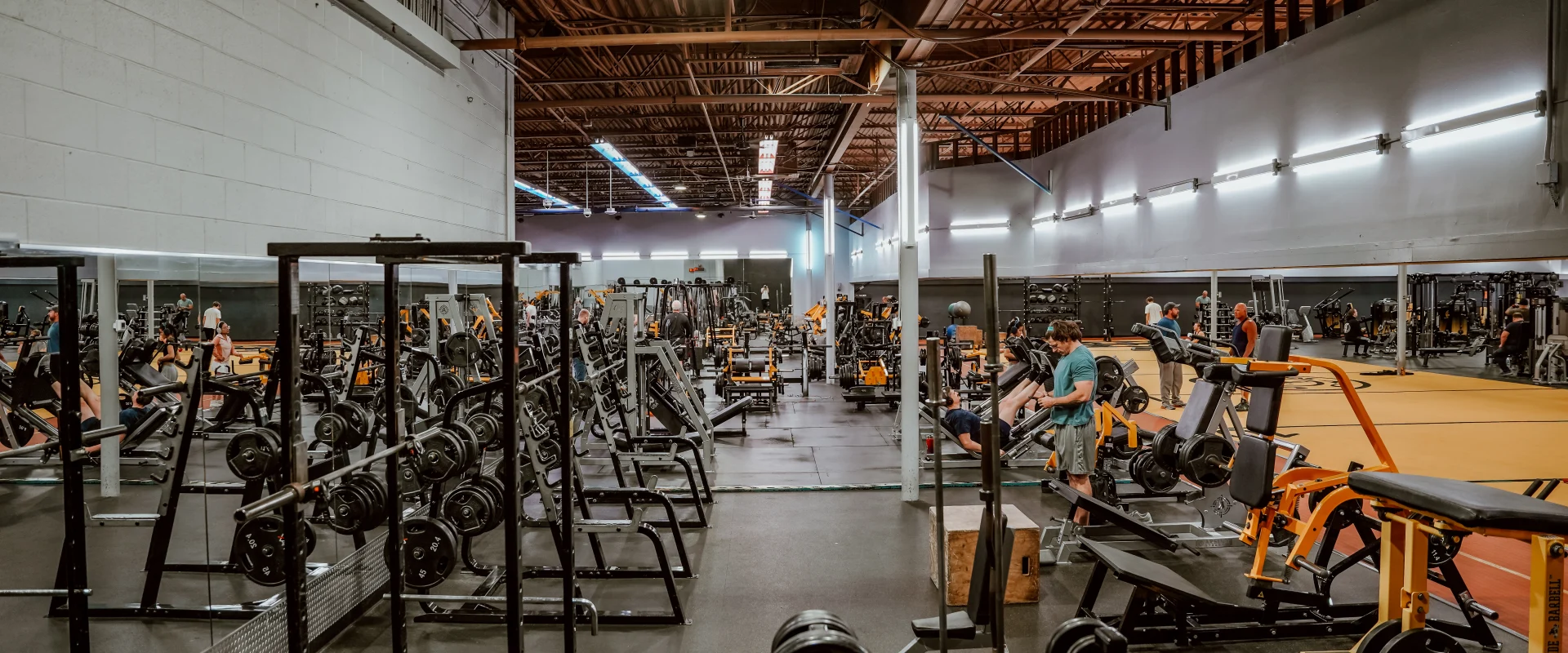 Plate-loaded equipment area at Gold's Gym Murfreesboro North, featuring a variety of weightlifting machines, barbells, and benches on a black rubber floor with a mirrored wall and an open industrial ceiling with warm lighting.
