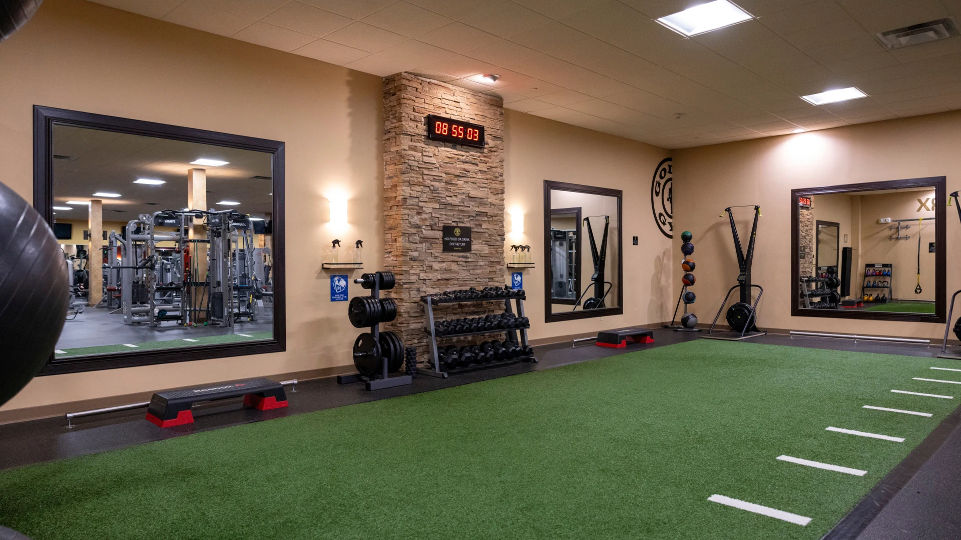 A functional training area in a gym with green turf flooring, white line markings, a stone accent wall with a digital clock, mirrors, dumbbell racks, and neatly arranged gym equipment like medicine balls, kettlebells, and resistance bands.