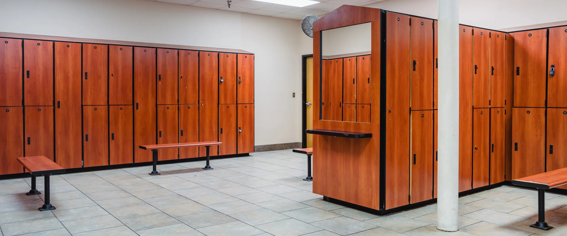 Locker room at Gold’s Gym in Austin (Southeast), featuring rows of wooden lockers with black trim and matching benches for seating. The space has tiled flooring and overhead lighting, creating a clean and organized environment.