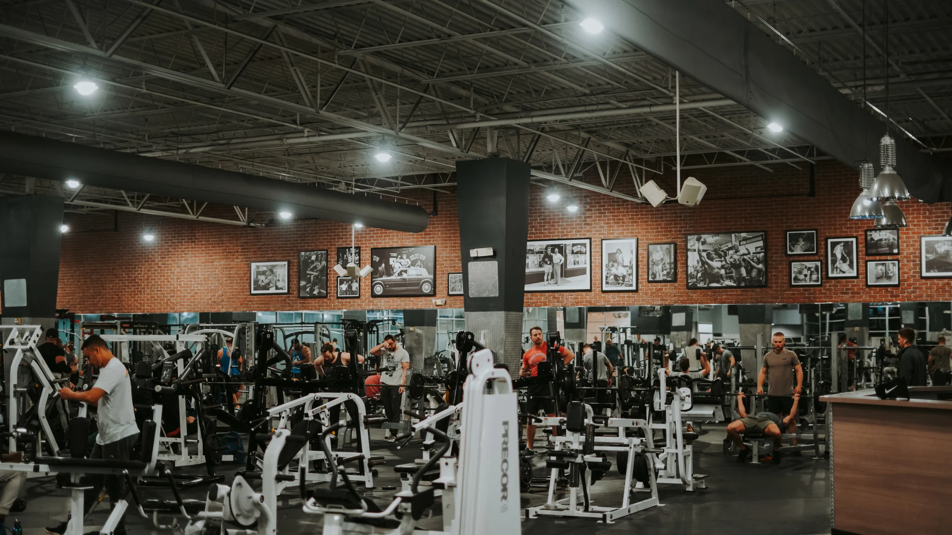 Machine weights area at Gold’s Gym Tennessee, Lee Highway, featuring rows of resistance machines and equipment arranged under a high ceiling with exposed beams and ducts. The brick accent wall in the background is adorned with framed historical photos, adding a classic touch to the space. Overhead lighting illuminates the area, while gym members actively engage in their workouts.