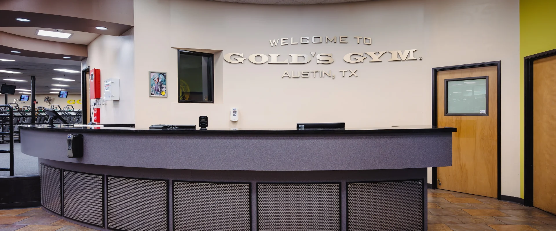 Front desk at Gold’s Gym in Austin (Southeast), featuring a wide, curved reception counter with dark finishes and a “Welcome to Gold’s Gym” sign on the wall. The tiled floor leads into the open gym space, with workout equipment visible in the background.