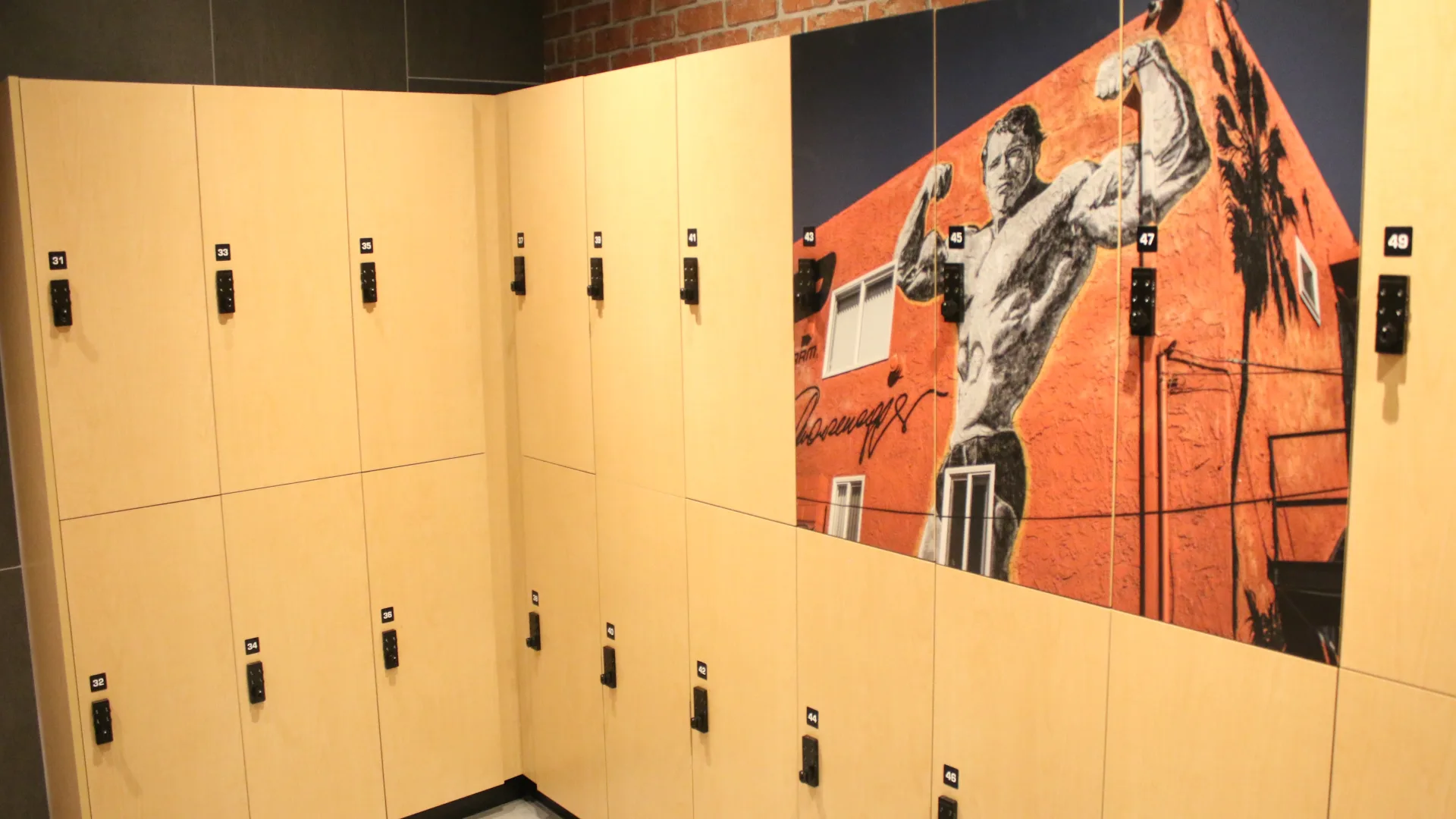 Light wood lockers in the Gold's Gym Austin, Burnet Rd locker room with a mural of a bodybuilder flexing on an orange building wall, adding a nostalgic, iconic gym feel.